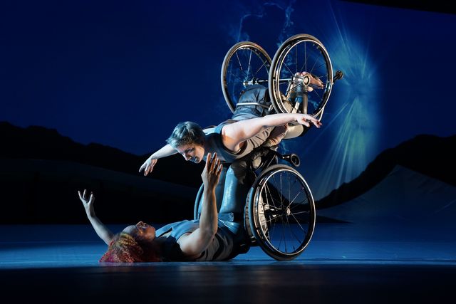 Laurel Lawson, a white dancer, balances on Alice Sheppard’s footplate, with arms spread wide, wheels spinning.  Alice, a multiracial Black woman with coffee-colored skin and curly hair, lifts Laurel from the ground below. A burst of white light appears in a dark blue sky.